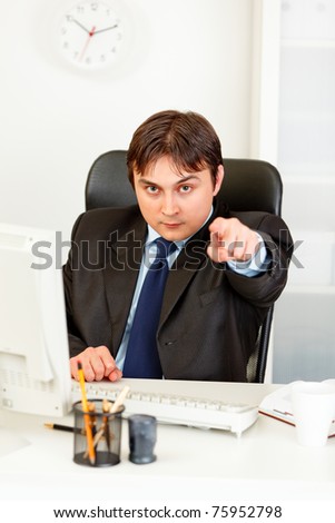 Confident modern businessman sitting at office desk and pointing finger at you