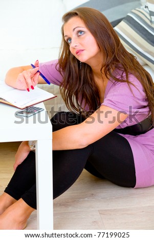 Thoughtful woman sitting on floor at home and making notes in diary
