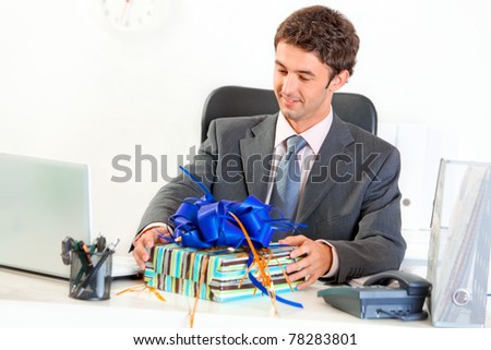 Pleased modern businessman sitting at office desk and looking on gift