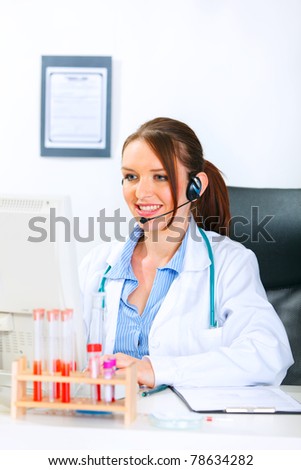Smiling medical doctor woman with headset sitting at office table and working on computer