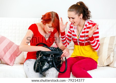 Indignant young girl sitting on sofa and looking on searching something in handbag girlfriend