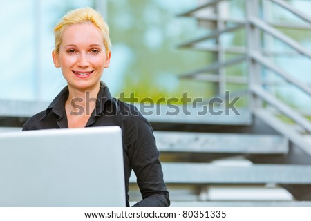 Pleased modern business woman sitting on stairs at office building and using laptop