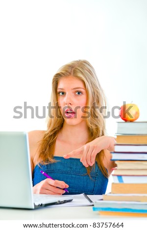 Curious young girl sitting at table with books and pointing on laptop