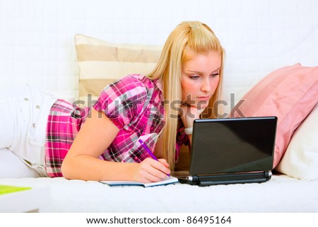 Concentrated pretty woman lying on couch with laptop and writing in notepad