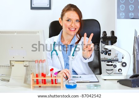 Smiling female doctor sitting at office table and showing victory gesture