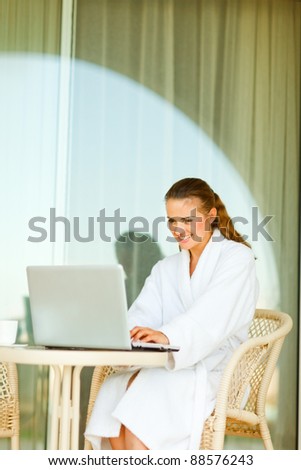 Smiling pretty woman in bathrobe sitting at table on terrace and using laptop