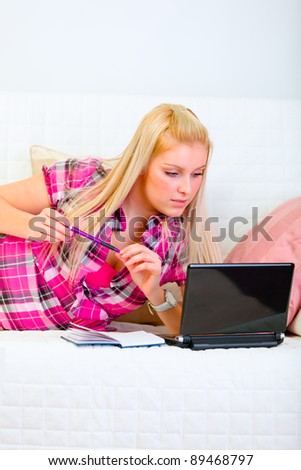 Pretty young woman laying on couch with notepad and pen and looking on laptop