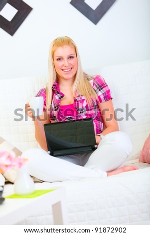 Smiling young woman sitting on couch with laptop and cup of coffee