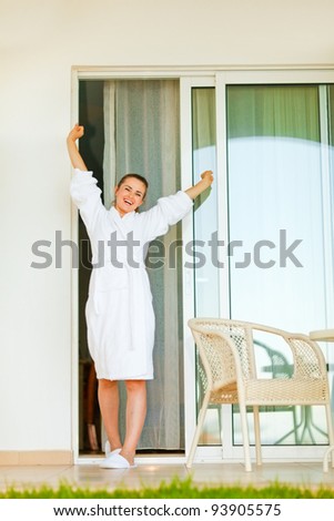 Happy beautiful female in bathrobe coming out on on terrace and stretching after sleeping