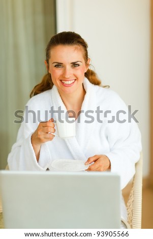 Portrait of smiling woman in bathrobe with cup of coffee and laptop on table