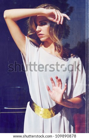 sad beautiful blond woman with curly long hair standing behind the window in long white dress and gold metallic belt