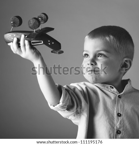 young 3 year old boy wearing long sleeve shirt playing with wooden toy airplane in his hand on studio background in black and white