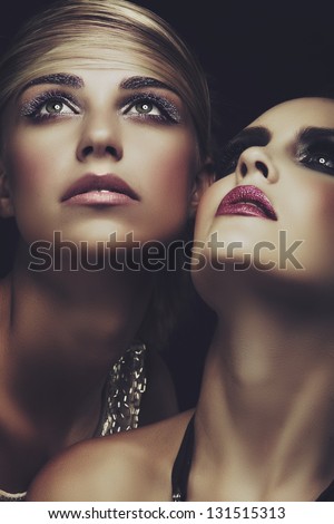 Closeup portrait of two beautiful young women with dark evening makeup on green eyes touching faces. Purple mascara, glitter on lips and eyes
