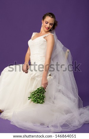 beautiful bride in ivory chiffon dress with lace and long tule veil sitting  against purple studio background