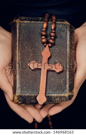 closeup of antique leather torn book with a christian cross in hands of a young woman held against black dress