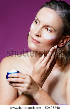 beautiful forty year old woman with natural makeup and healthy skin texture on pink studio background. Applying facial cream from a cosmetics tub