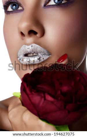Closeup of woman\'s face with silver lipstick. Blue contact lenses, purple eye shadows and tanned hand with red metallic and texture manicure holding red fabric flower