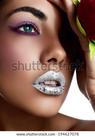 Closeup of woman's face with silver lipstick. Blue contact lenses, purple eye shadows and tanned hand with red metallic and texture manicure holding red fabric flower