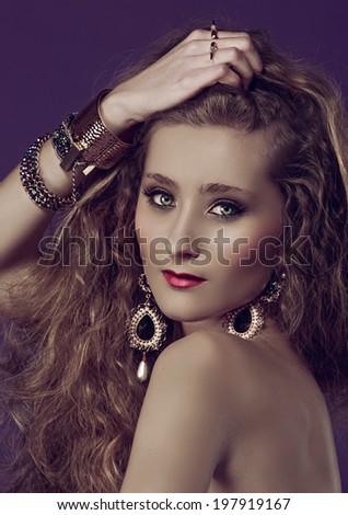 Portrait of a young beautiful woman with long blond curly hair wearing pearl earrings and gold bracelets against purple studio background