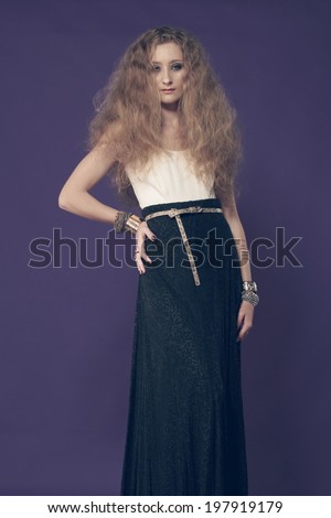 Portrait of a young beautiful woman with long blond curly hair wearing pearl earrings and gold bracelets, long black skirt against purple studio background