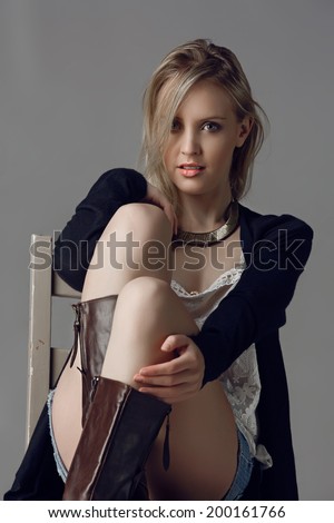 Beautiful blond woman wearing black cardigan, jean shorts and leather boots on grey studio background sitting on a rustic chair