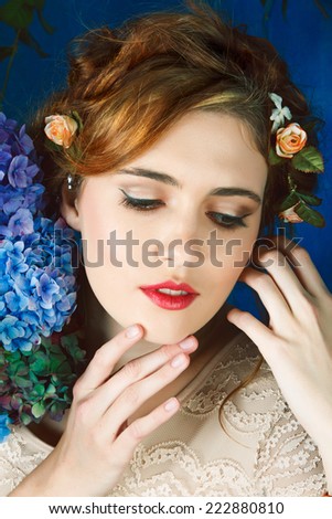 Romantic portrait of a beautiful woman with red hair and flowers in her hairstyle, with hand and flowers touching her face against blue grunge background