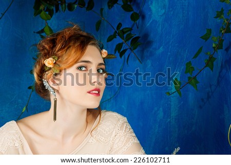 Romantic portrait of a beautiful woman with red hair and flowers in her hairstyle, wearing diamond ear cuff against blue grunge background