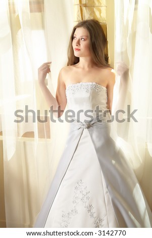 Beautiful young bride with long brown hair in white and silver satin dress at window, natural light