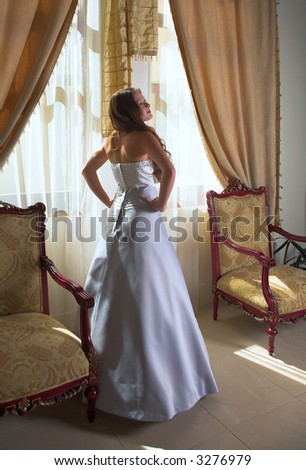 Beautiful bride in silk grey and white dress standing next to sunlit window with heavy curtains