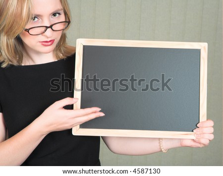 Young serious blond woman teacher in black dress and pearls, wearing glasses pointing at empty blackboard