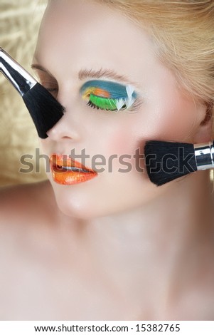 Blond woman with green fantasy long lashes and bright make-up during make-up application with brushes touching her face