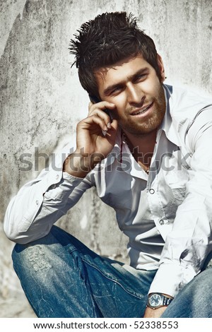 handsome fashion model in white cotton shirt and blue rugged jeans talking on the cell against grunge wall.