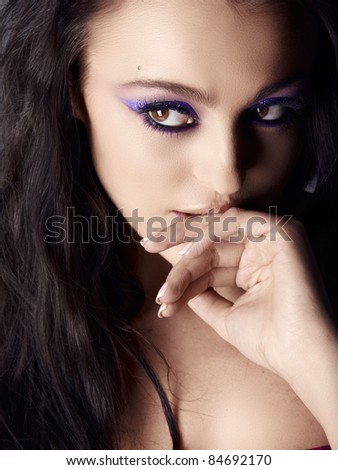 Beautiful young Italian woman with artistic purple eyeshadow and long hair looking away and biting her hand, shot in low key