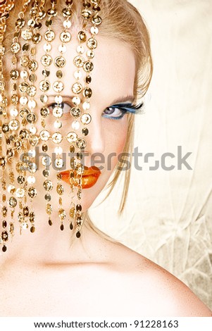 Close-up portrait of a sensual beautiful blond woman model face with bright fashion make-up, orange lips and long eyelashes hiding behind gold accessories