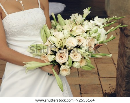close-up of a bride wearing white dress holding pale cream roses and green lilies bouquet on a summer day outdoors.