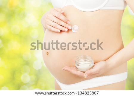 Pregnant woman applying skin care cream on belly over green fresh background. Prenatal health concept