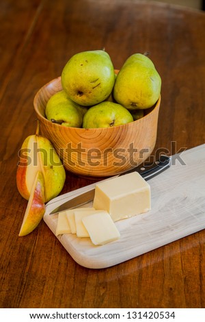 A bowl of fresh Bartlett pears on a wood table with sliced cheese and a paring knife on a wood cutting board and one cut pear
