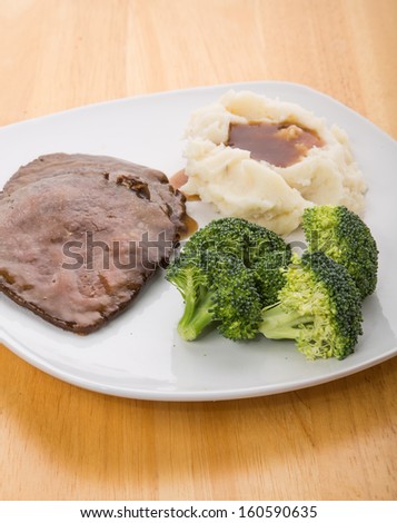 A dinner of sliced roast beef, mashed potatoes with gravy and fresh broccoli on a white plate on wood table.