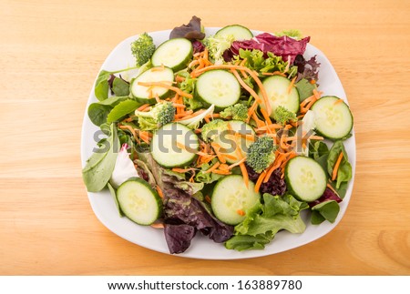 Salad of mixed greens, broccoli, cucumber slices, shredded carrots in a white plate on a wood table.