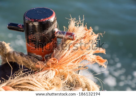 Frayed nautical rope tied to an old rusty cleat with water in background