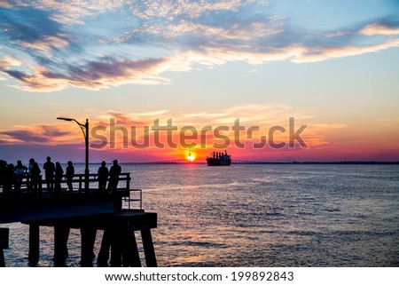 A large, empty freighter sailing toward a suspension bridge at sunset with People on Pier Sillouetted