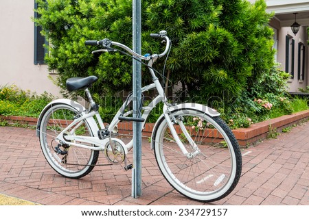 An old white bicycle locked to a sign post on a brick sidewalk in front of a green shrub and home