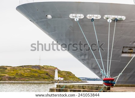 Bow of a massive white cruise ship moored to red bollard with blue ropes with white lighthouse in background