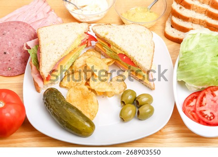 A delicious ham, cheese, lettuce and tomato sandwich with mustard and mayo on a plate with a dill pickle, potato chips and olives, with ingredients on wood cutting board