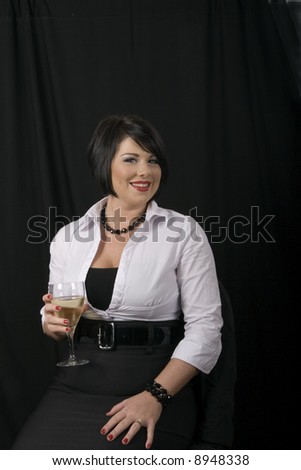 A black haired woman in white blouse against a black background sitting in a chair with a glass of wine smiling at the camera