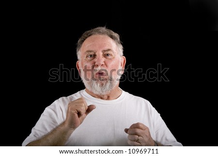 An older man in a white t-shirt on a black background looking as if he forgot or lost something
