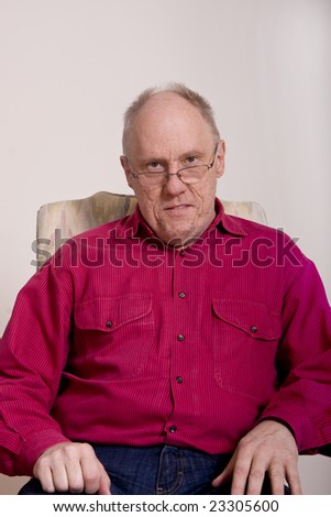 An older bald guy in a red shirt sitting in a chair wearing glasses