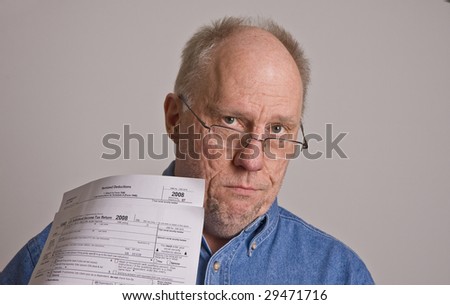 An older bald man in a blue denim shirt on a grey background wearing glasses and holding tax forms