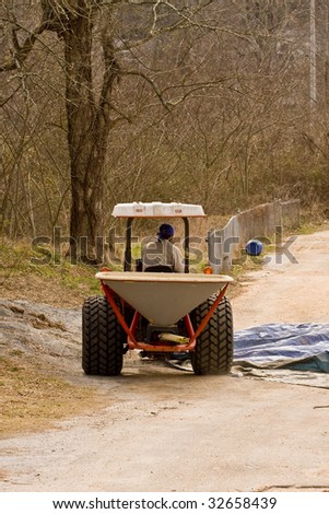 A worker driving a seed spreading machine down a dirt road