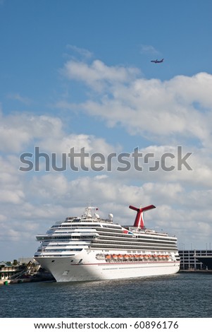 A large luxury cruise ship in a harbor with a commerical airline flying overhead